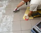 Pracownik w rękawiczkach nakłada fugę epoksydową na płytki podłogowe za pomocą kielni. Proces fugowania krok po kroku.