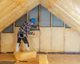 Pracownik w kasku izoluje strych, który jest kondygnacją, wełną mineralną.
